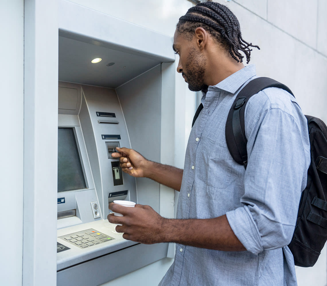 A man wearing a blue collared shirt with a black backpack is standing at an ATM machine, withdrawing cash in a well-lit urban setting.